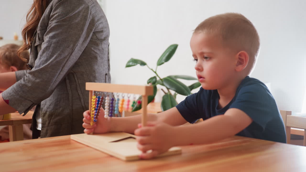 Niño pequeño vestido de azul mueve cuentas en un ábaco de madera mientras un compañero toca el marco; una mujer se gira para ayudar a otro niño; escena tranquila de preescolar centrada en la motricidad fina y la concentración.