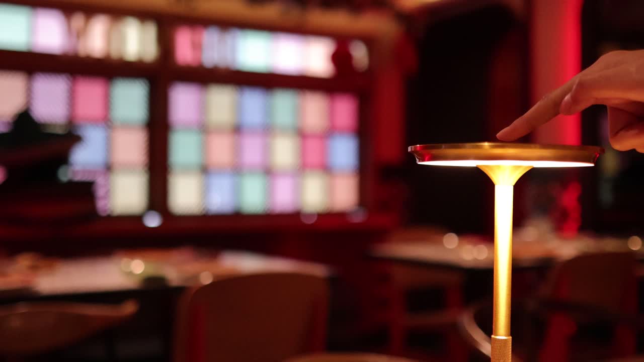 A hand reaches out to turn on a sleek, modern touch lamp in a warmly lit, colorful interior with blurred background and soft ambient lighting