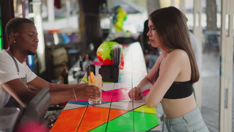 barman treats woman with cocktail at bar counter. black barkeeper puts straw into drink and gives glass to guest in club. bartender serves visitor