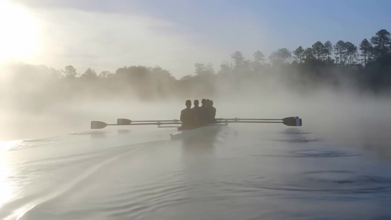 A serene rowing team glides through a foggy river, surrounded by lush trees and calm water, creating a peaceful and natural setting for their sport. Front view of the rowers with wooden oar.