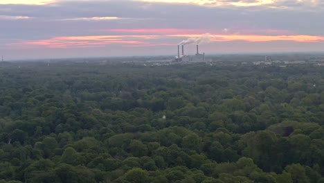 Drone-shot-backwards-of-a-power-plant-at-the-wide-horizon-over-green-trees-of-Munichs-Englischer-Garten-including-a-lake