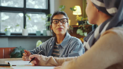 Muslim and Hispanic Female Students Talking at Desk in Classroom