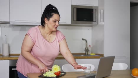 Young-overweight-woman-cooking-salad-at-kitchen,-watching-video-recipe-on-laptop-online