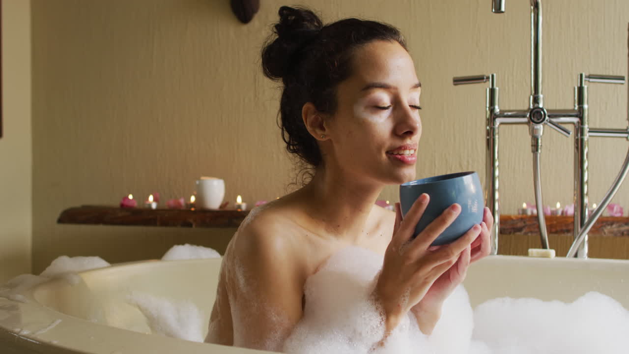 Relaxed biracial woman lying in bath with foam and drinking coffee