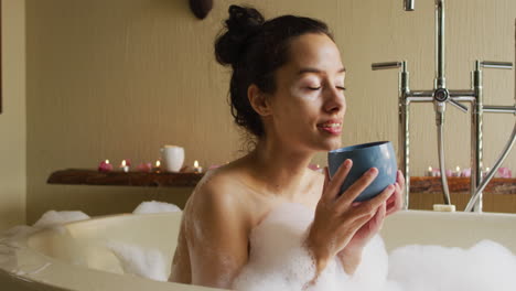 Mujer-Birracial-Relajada-Tumbada-En-El-Baño-Con-Espuma-Y-Bebiendo-Café