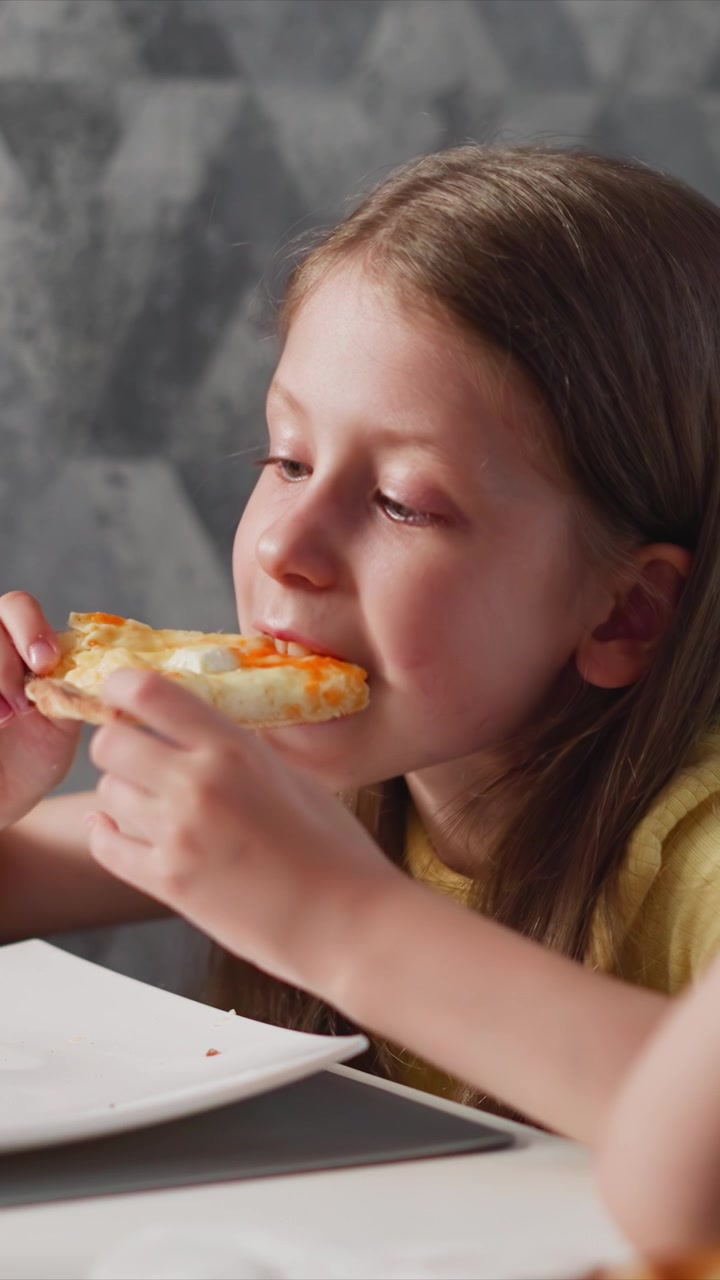 Close up of young girl eating pizza. Girl holds slice takes bite from plate on table with gray geometric background. Soft lighting highlights her focused expression as girl enjoys meal