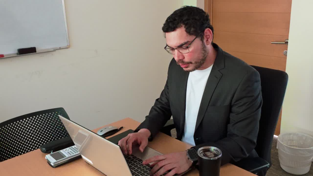 A man wearing glasses is sitting at a desk with a laptop and a cup of coffee