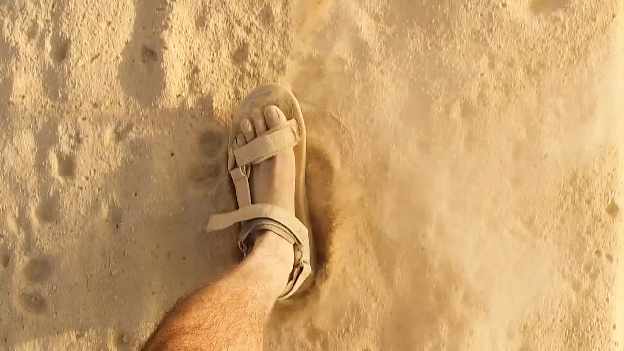 hombre caucásico de primer plano con pies polvorientos caminando a cámara lenta con sandalias en el desierto arenoso o en la playa, huellas pateando algunas partículas polvo brillante arena usando sandalias en un día soleado