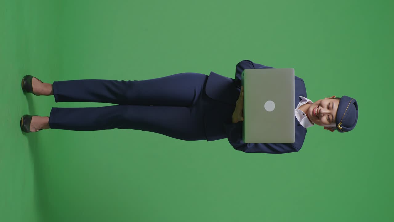 cuerpo lleno de mujer asiática azafata sonriendo y usando una computadora portátil mientras está de pie en el estudio de fondo de la pantalla verde