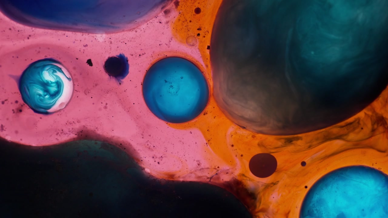 Microscopic blue and teal spheres moving through a vibrant orange liquid. Close-up shot showing chemical reactions and artistic fluid dynamics.