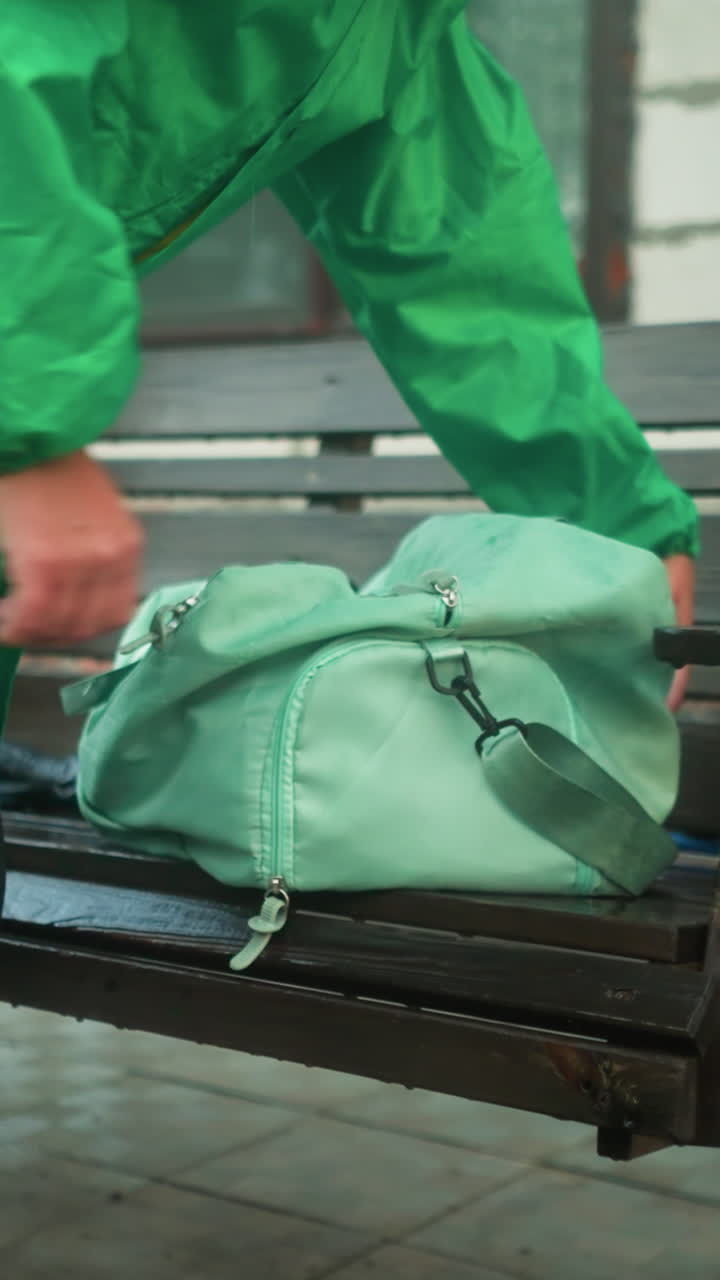 primer plano de un individuo con una impermeable verde y leggings alcanzando una bolsa verde menta en un banco oscilante, la escena incluye un entorno al aire libre con un banco, una cadena y un edificio en el fondo
