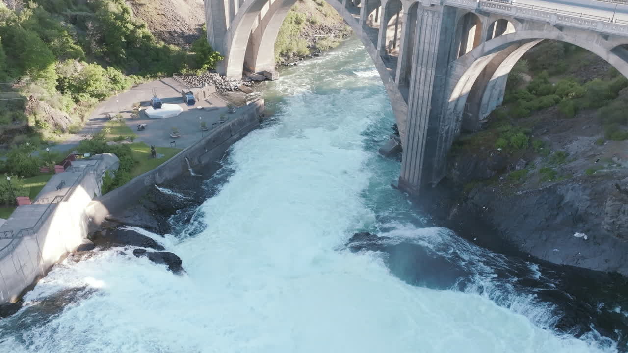 Whitewater rushes under a concrete bridge. You can see park structures and cables on the riverbank