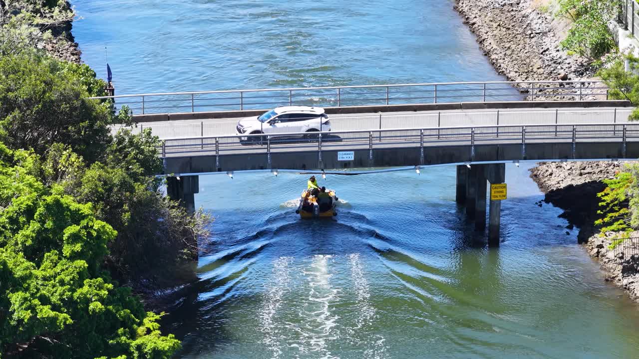 Two workers pilot a small barge beneath a canal bridge, aerial view, bright daylight