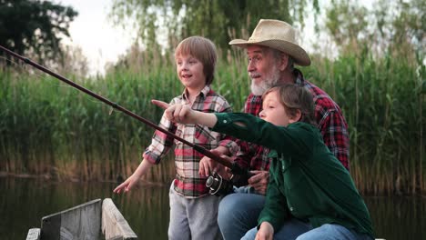 cute teen boys showing something to their grandfather while they are fishing sitting on the lake pier