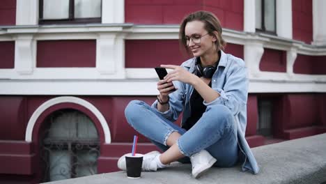 A-Fashionable-Girl-In-Glasses-Sits-On-The-Street-On-A-Parapet,-Concentrates-On-Her-Mobile-Phone