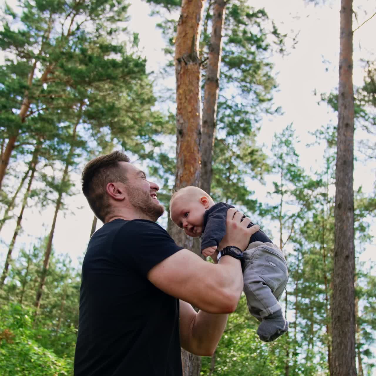 Handsome muscular Caucasian man tossing his little baby boy. Spending time with child in nature. Low angle view. Pine trees at backdrop