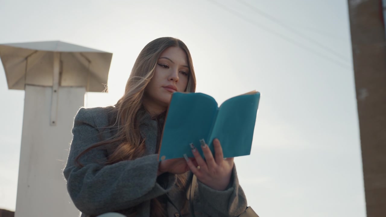 Donna illuminata dal sole con capelli fluenti che studia all'aperto. Donna elegante sul tetto che si gode l'alba leggendo. Signora elegante con un libro turchese tra la luce del sole e i capelli mossi dal vento all'aperto.