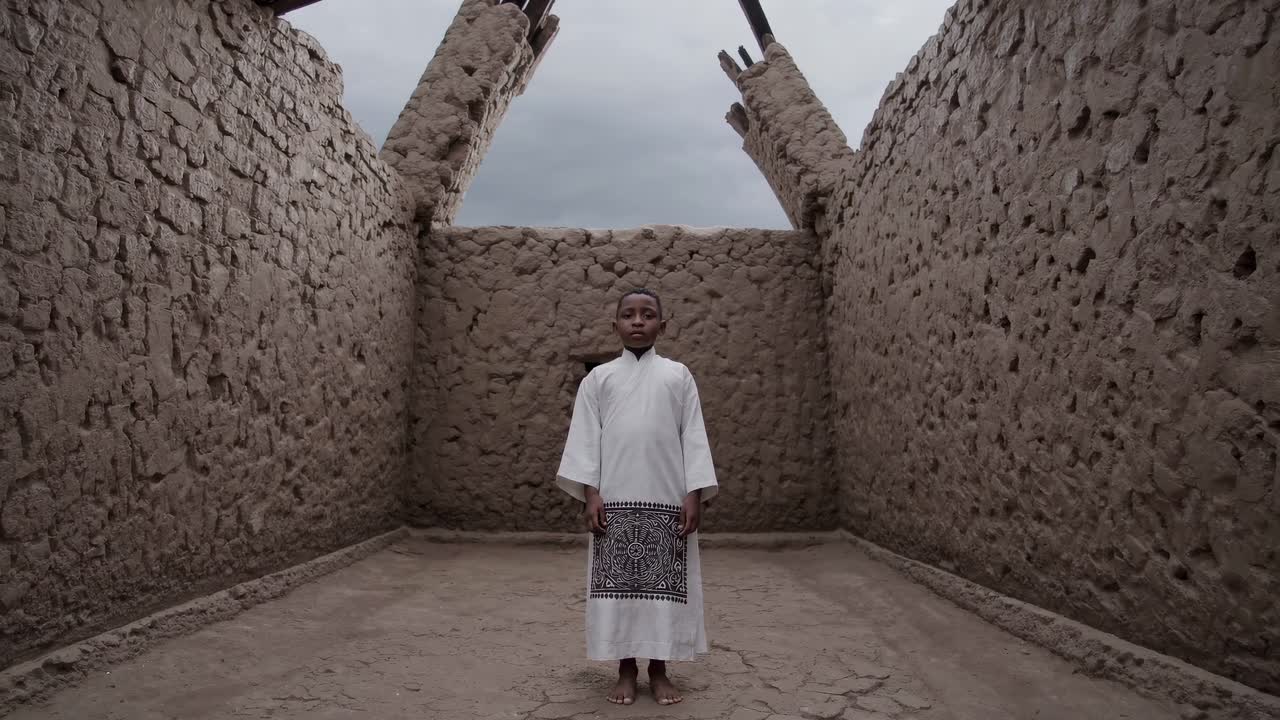 Young boy stands confidently in a rustic room with earthen walls, showcasing a moment of contemplation and connection to his surroundings in a serene atmosphere