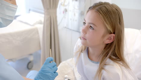 Caucasian-female-doctor-in-mask-and-gloves-checking-throat-of-girl-patient-in-hospital,-slow-motion