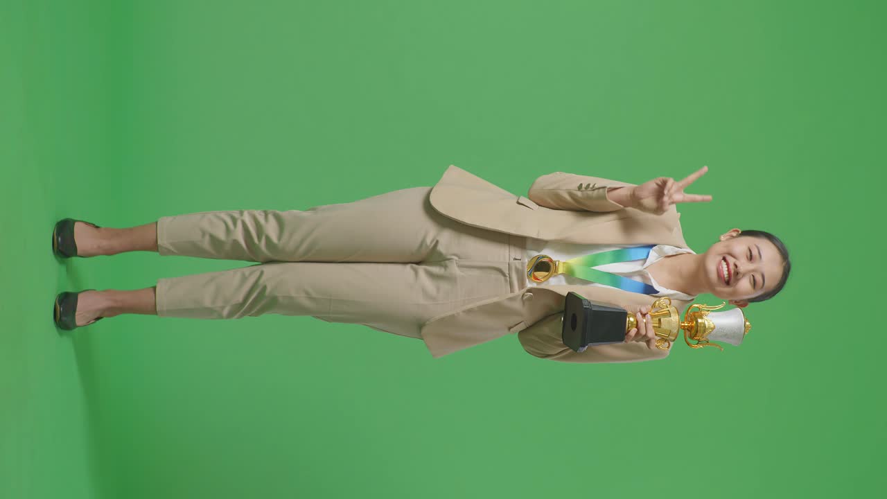 금메달과 트로피를 입은 슈트를 입은 아시아 사업가 여성의 몸이 평화의 몸짓을 보여주고 스튜디오의 녹색 스크린 배경에서 첫 번째 우승자로 카메라에 미소 짓고 있습니다.