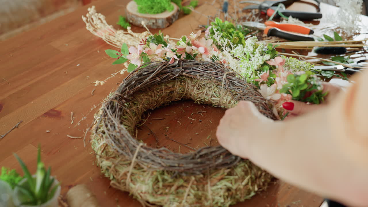 Hands of florist decorating straw and twig wreath with fresh greenery and pink blossoms on wooden table, surrounded by tools, dried flowers in creative floral workshop arrangement