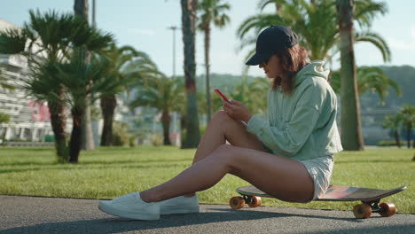 woman sitting on a skateboard in a park using a phone