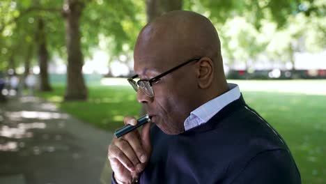 professional bald black male vaping while sitting outdoors on a bench in a park