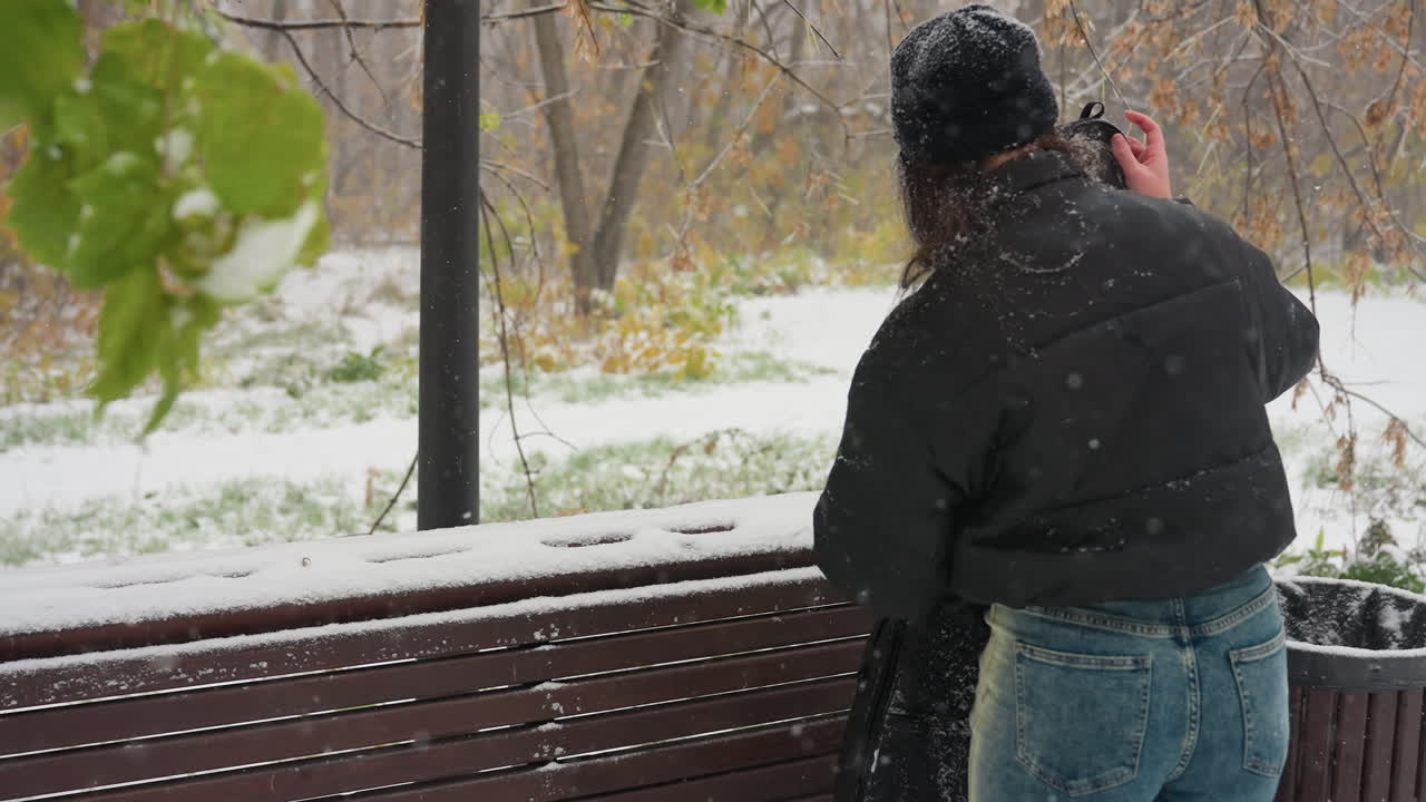 냉정 한 겨울 공원 에서 눈 으로 인 의자 에 놓인 여자 가 기타  을 열고 있는 모습, 눈 이 부드럽게 떨어지고, 초록색 과 얼어붙은 나무 들 이 여 있는 모습