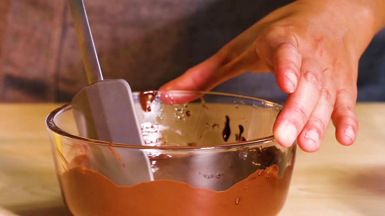Hands using a spatula to mix chocolate ganache in a clear glass bowl on a wooden surface.