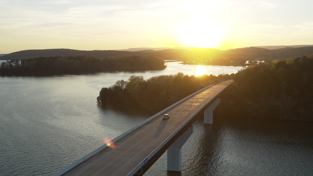 Riprese aeree che seguono un'auto mentre attraversa un ponte su un lago con il tramonto sullo sfondo.