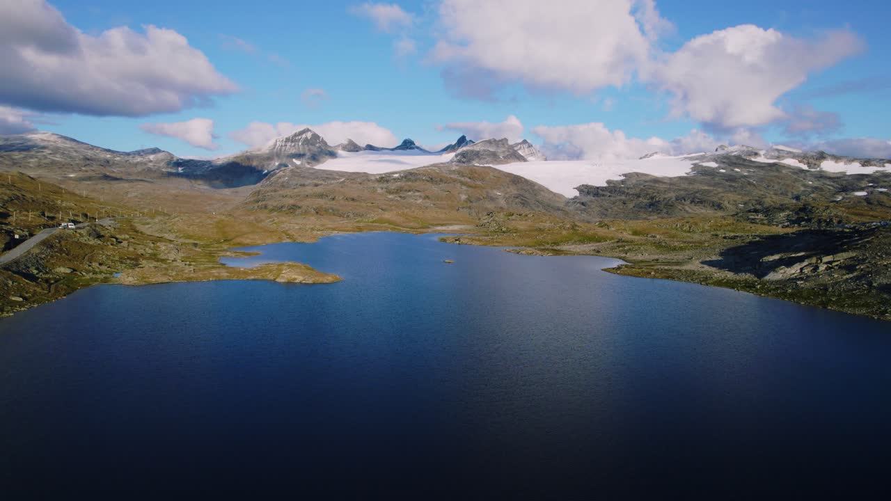 Panoramic view over still lake water surrounded by mountains, glaciers and rocks and sparse vegetation in Norway