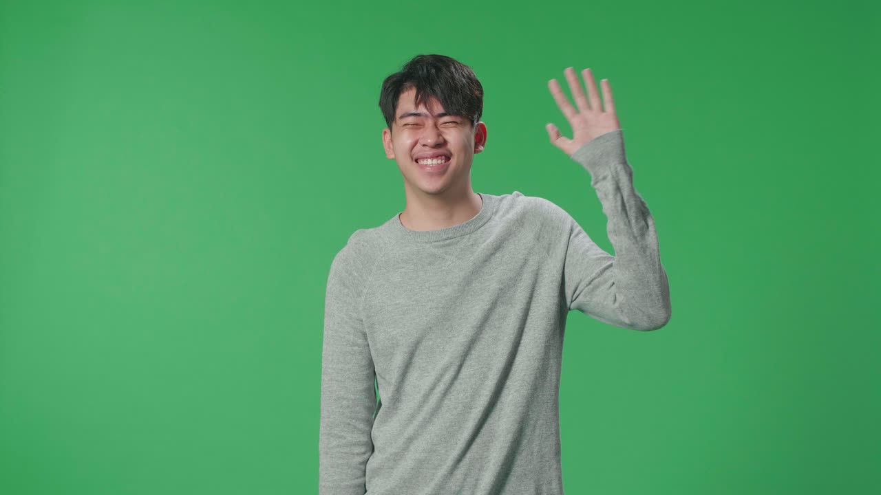 un hombre asiático agitando la mano y sonriendo mientras está de pie en la pantalla verde en el estudio