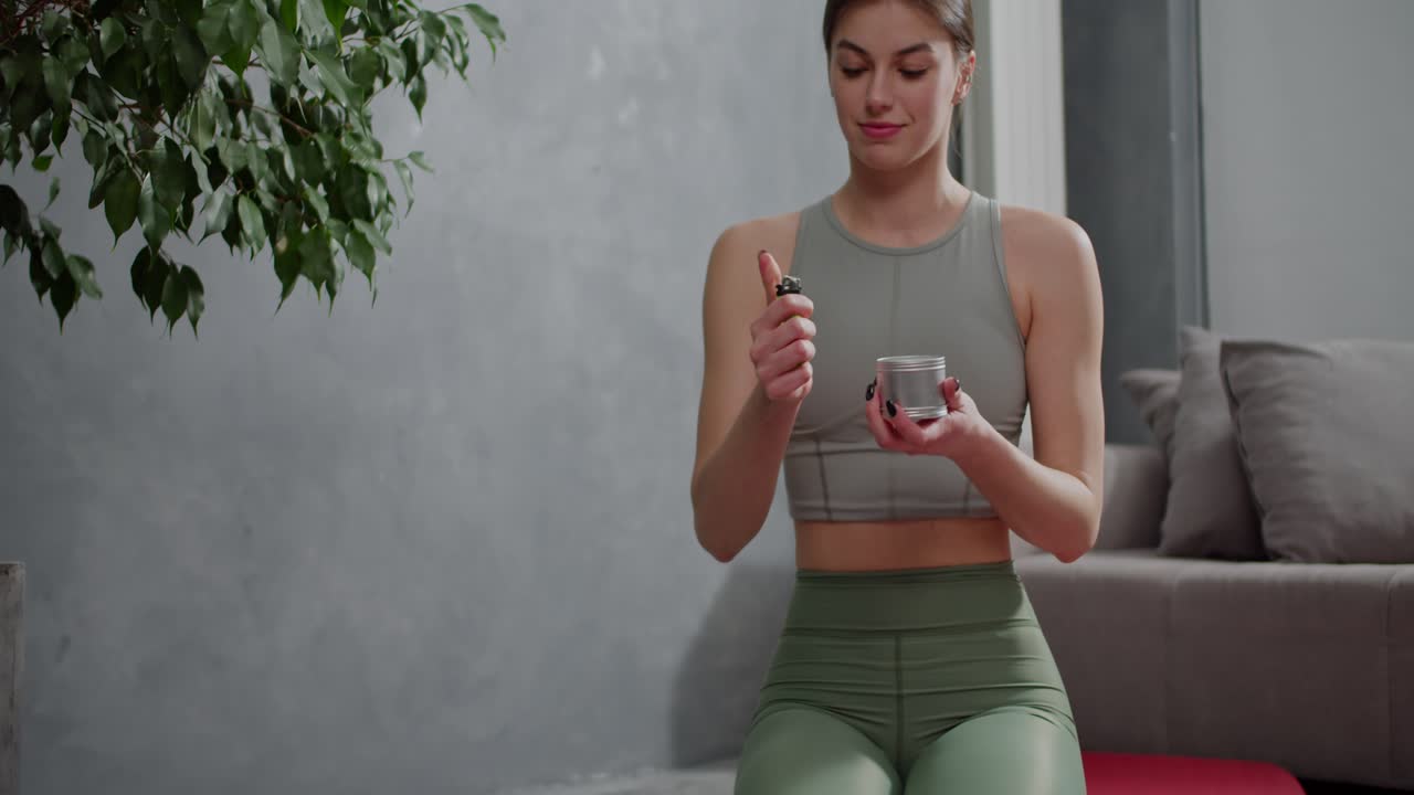 tranquila y segura chica morena en un uniforme deportivo de verano con una camiseta gris y pantalones verdes enciende una vela casera antes de su clase de deportes y yoga en un apartamento moderno en casa