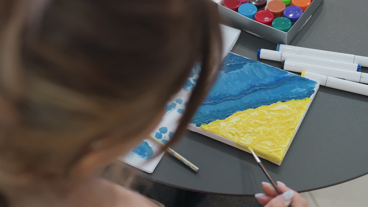 Pintura artística costera, artista realzando meticulosamente una obra de arte con textura azul y amarilla del océano, artista blanca concentrada cuidadosamente en un paisaje marino con textura azul y amarilla sobre lienzo