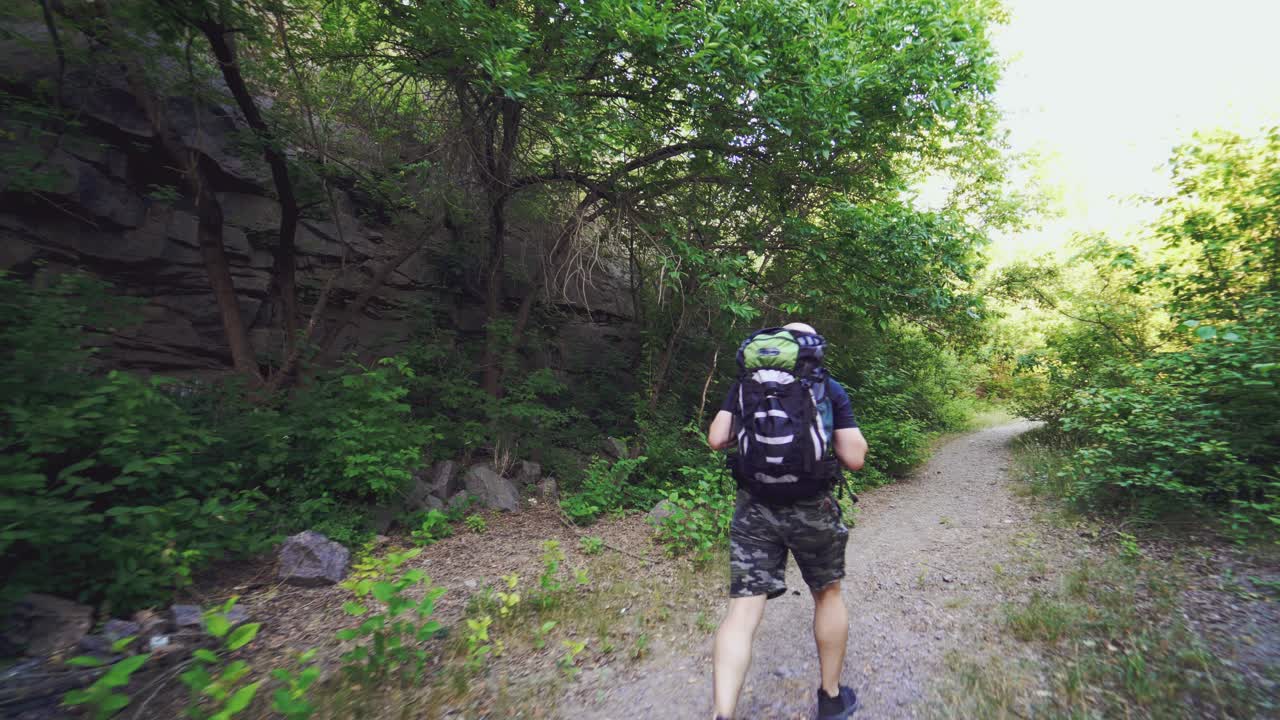 A man with a backpack on his shoulders is climbing up the path against the background of green trees in the summer on a warm day. Tourist in a hike.