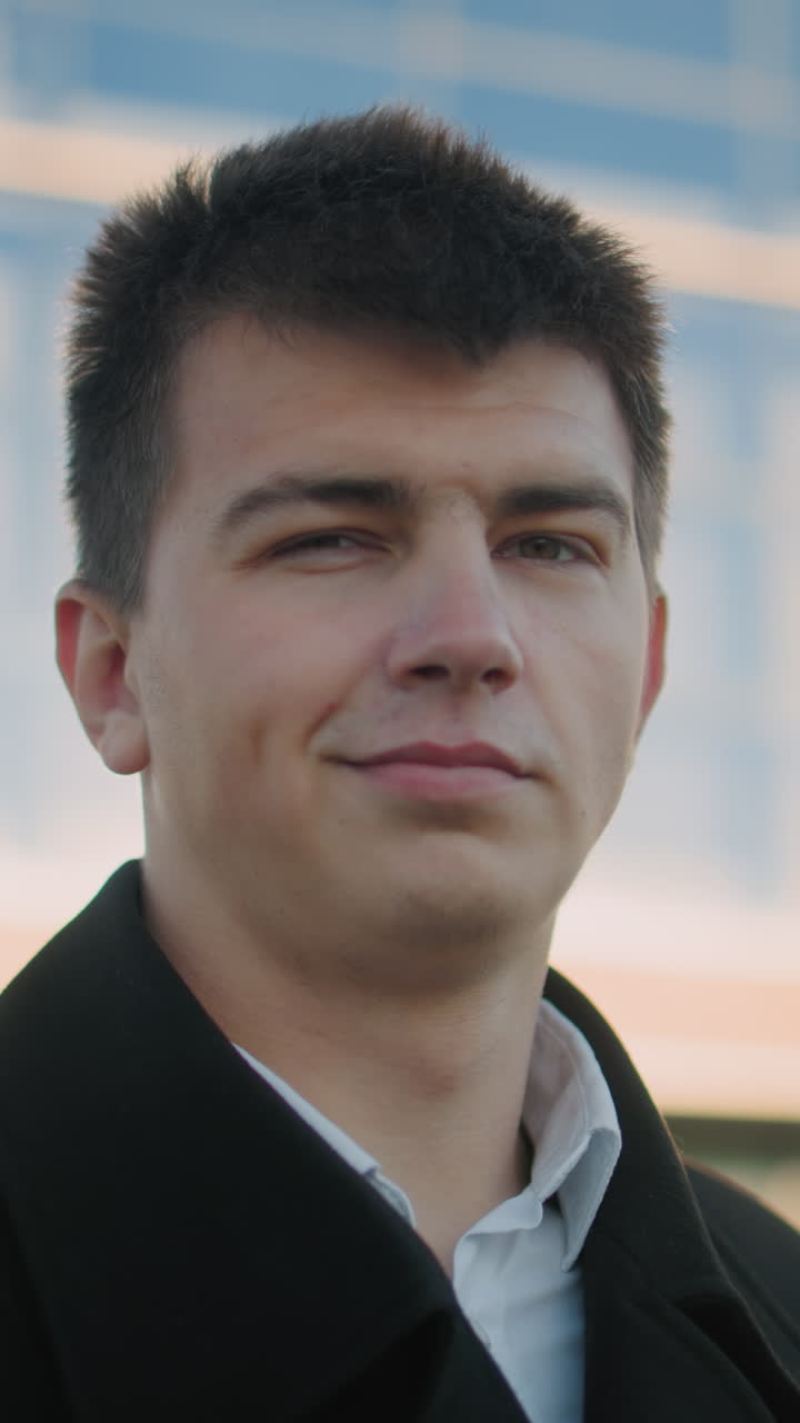 검은 옷을 입은 남자가 카메라를 향해 자신감 있게 미소 짓고, 뒤에서 부드러운 밝은 빛이 비추고, 배경에는 반사 유리 건물이 있습니다.