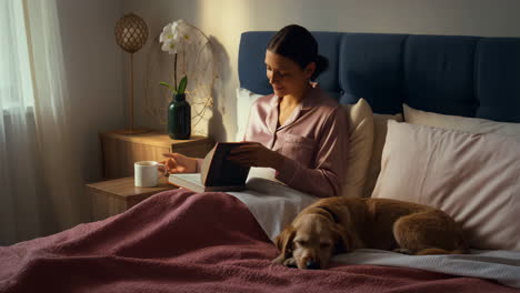 woman reading and drinking coffee in bed with her dog