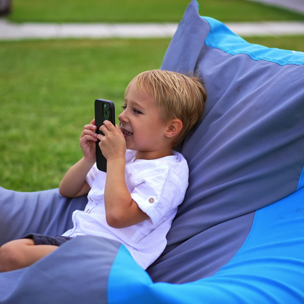 Nice cute kid sitting outdoors in comfortable chair. Blond boy speaking on the smartphone on loud speaker. Green grass at backdrop