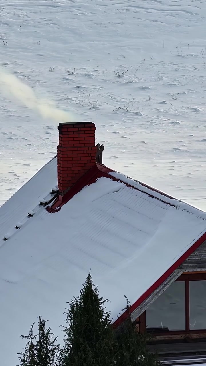 Close up of a red brick chimney with smoke blowing in the wind on a snowy rooftop. Ideal for winter themes, home heating, and cozy mountain living concepts.