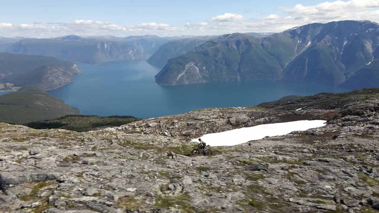 espectacular toma de dron que muestra a un ciclista de montaña montando en la cumbre rocosa de las montañas noruegas - hermoso valle con fiordo azul en verano