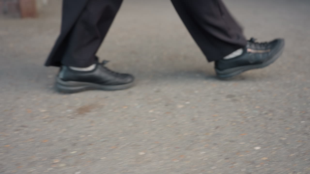 로우 앵글 트래킹 숏으로 검은 신발을 신은 사람이 거친 보도를 성큼성큼 걸어가면서 속도와 도시 통근의 에너지를 전달하고, 옷자락은 포장 도로 근처에서 찰랑거리고 꾸준한 발걸음은 거친 텍스처를 가로지르는 모습을 보여줘요.