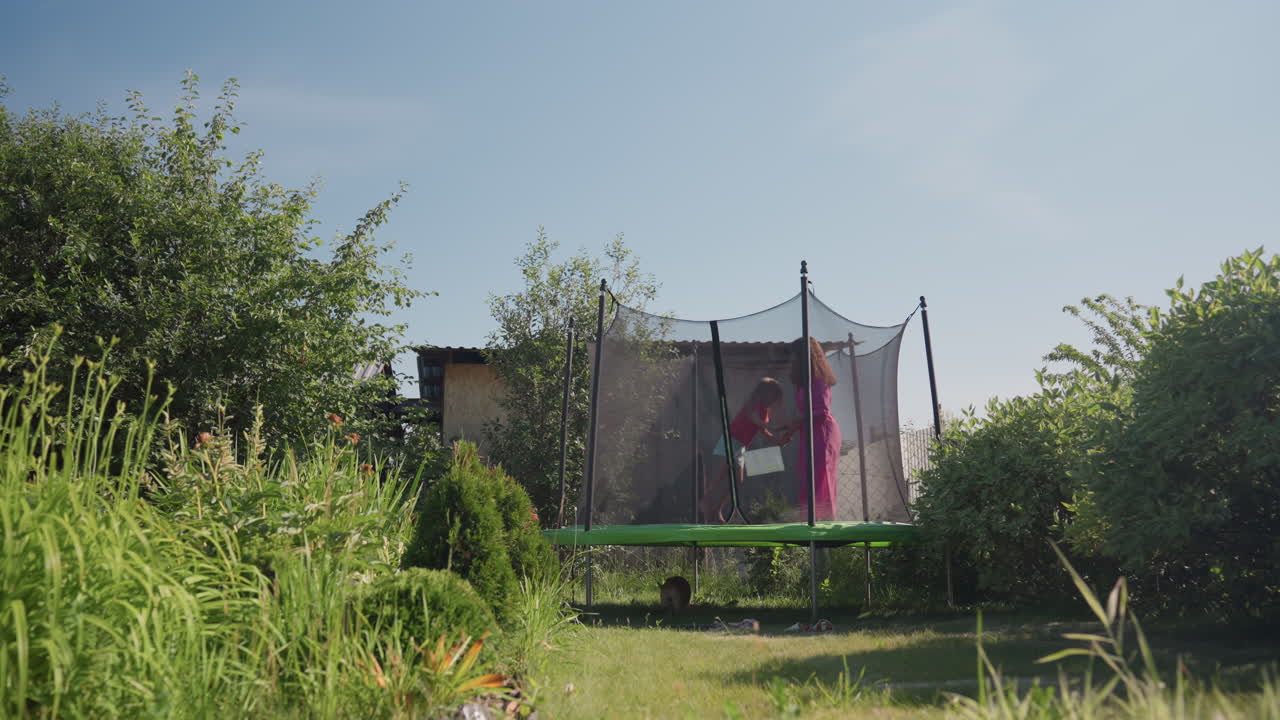 Girl Has Fun Outdoors, Young Girl Enthusiastically Plays On Springy Trampoline Among Green Leaves, Excited Child Happily Leaps On Trampoline Surrounded By Lush Leafy Trees And Plants