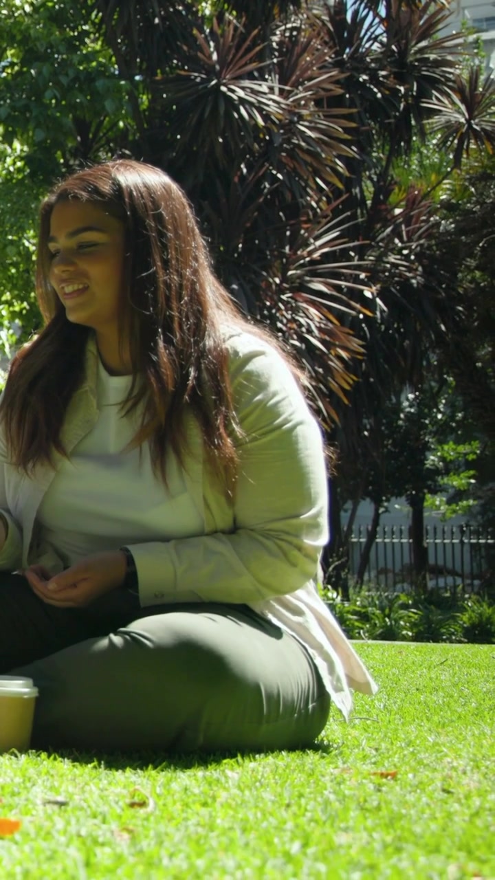 Video vertical: Palomas acercándose a una mujer con chaqueta y pantalones verde oliva, que ofrece comida en un parque con una taza.