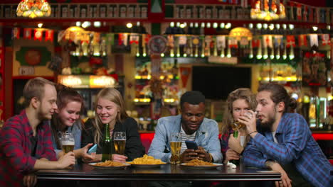 estudiantes alegres sentados en una mesa en un bar bebiendo cerveza comiendo papas fritas y viendo fotos en una pantalla de teléfono inteligente discutiendo fotos.
