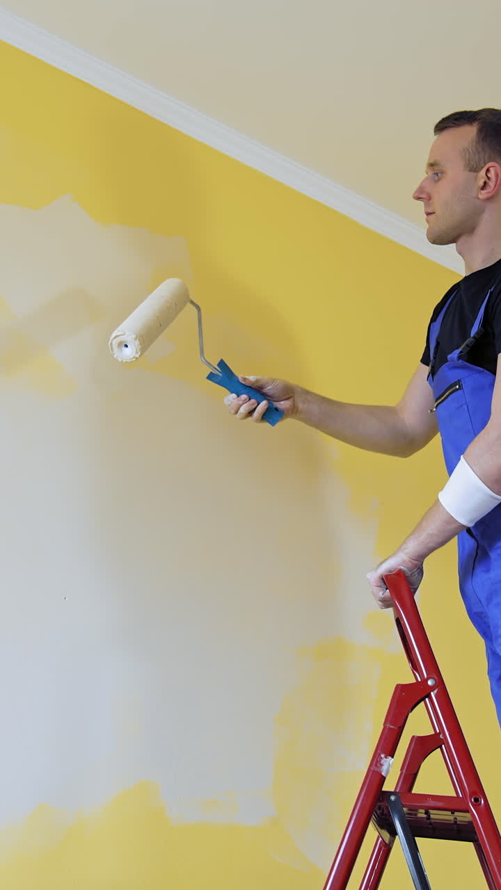 Worker repainting walls of the apartment from the ladder. Young builder male decorating walls into a new light color. Home repair. Vertical video