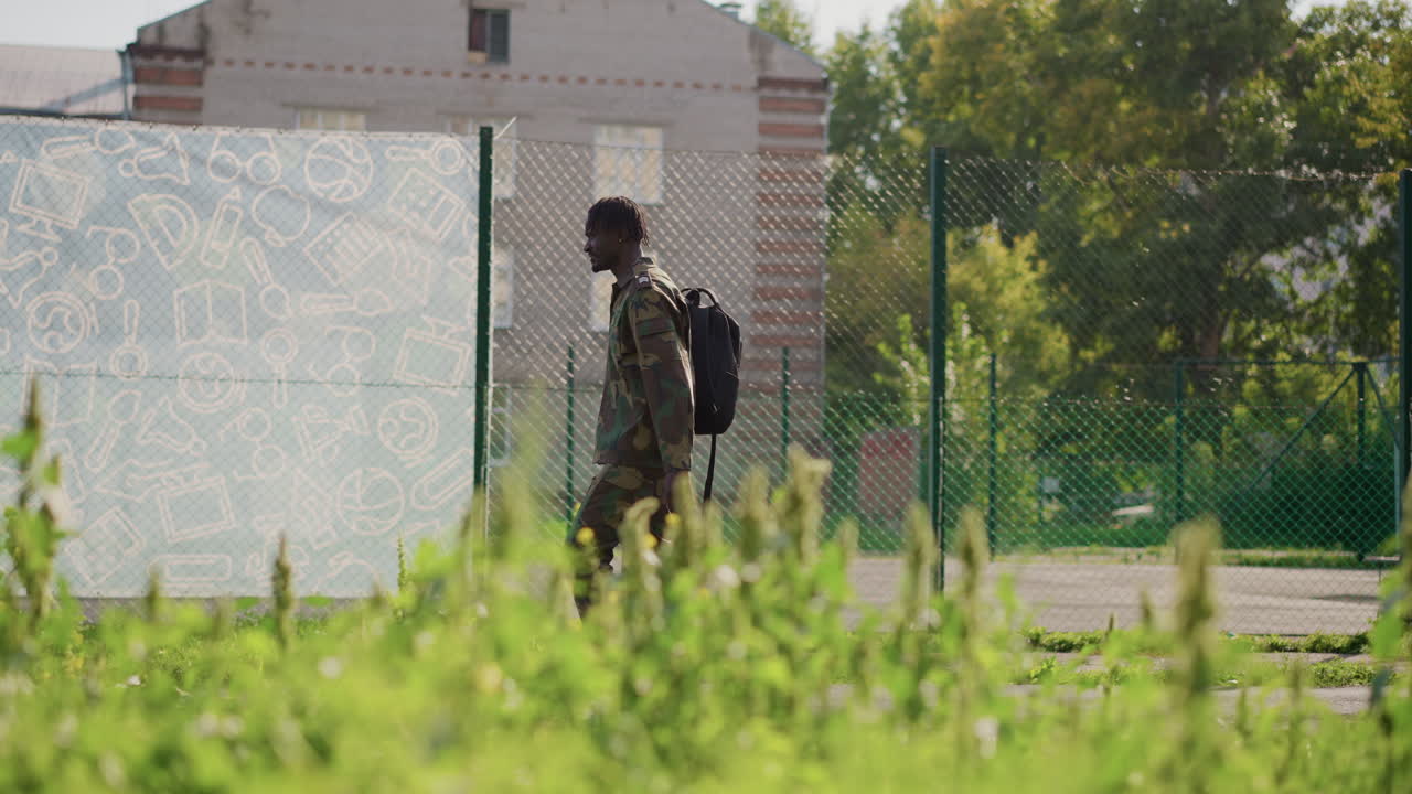 Black Former Soldier Walking Grassy Court With Backpack And Camouflage Jacket, Suburban Buildings And Sunlight Beyond. Quiet Stride And Downcast Gaze Reflect Reintegration And Healing After Service