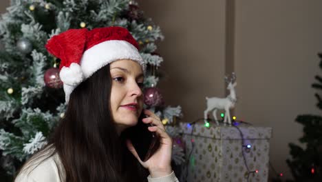 Hermosa-Morena-Con-Gorro-De-Papá-Noel-Habla-Por-Teléfono-Cerca-Del-árbol-De-Navidad-Y-Regalos