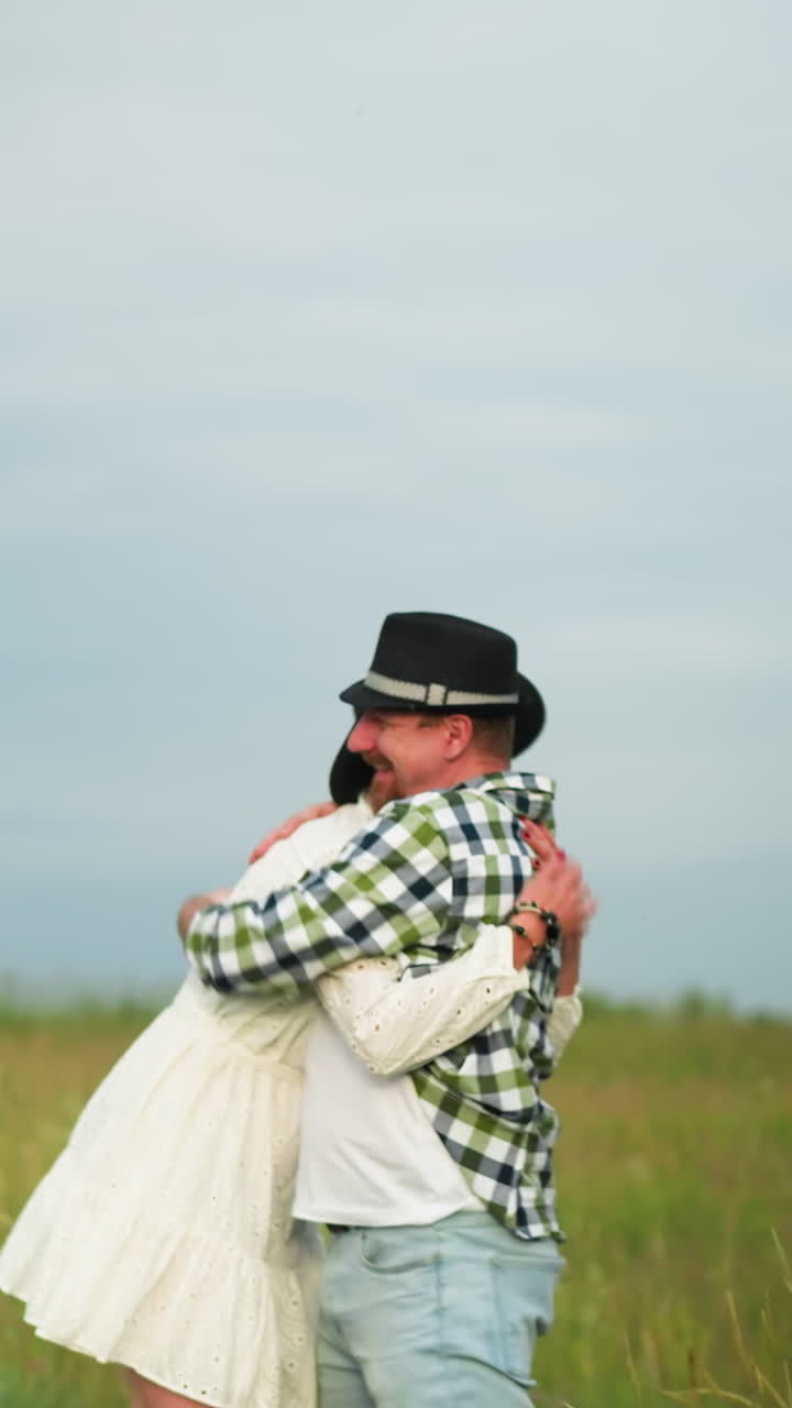 un hombre con un sombrero negro y una camisa a cuadros persigue a una mujer con un vestido y sombrero blancos a través de un campo cubierto de hierba bajo un cielo nublado. la escena concluye con un abrazo sincero
