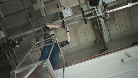 man wearing glasses and striped sweater stands on scaffold platform adjusting overhead track lighting in studio grid, surrounded by cables and fixtures, technical crew preparing broadcast set lighting environment