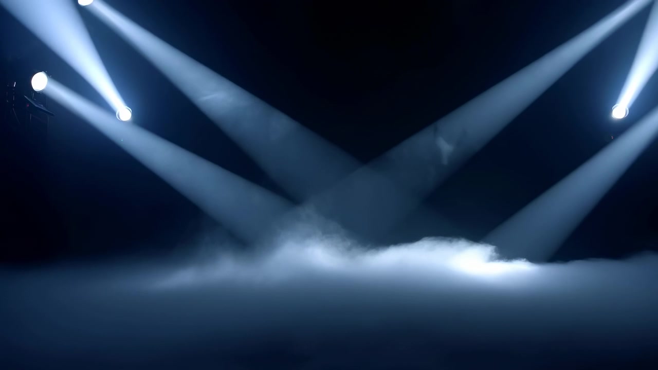 Moving beams of cool white light change position from side to side while dense mist spreads across the floor, creating layered illumination and strong contrast against a black background.